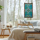 26"H Tiffany Style Stained Glass Brandi's Window Panel hangs on a window in a bright, modern bedroom. The scene includes a bed with a cream knit blanket, a wooden tray table, and a bench, with bare trees visible outside the window.