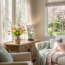 15.5" H Yara Multi Colored Stained Glass Vase-Shaped Table Lamp in a living room with a wooden table and floral arrangement, near a window with cherry blossoms outside.