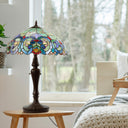 24.5" H Marlowe Multicolor Tiffany-Style Stained Glass Table Lamp sits on a small wooden side table in a bright bedroom setting. The lampshade features an intricate Tiffany-style mosaic design with blue, green, yellow, and red floral and bird motifs, set against a softly blurred window view of trees.
