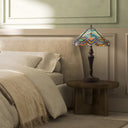24" H Highclere Victorian Multicolor Tiffany-Style Stained Glass Table Lamp sits on a round wooden nightstand next to a bed in a softly lit bedroom.