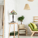 21.5" H Crimson Cardinal Tiffany-Style Stained Glass Cone Shade Candlestick Table Lamp sits on a light wood side table in a bright, modern room with a white brick wall, hanging woven pendant lights, a wooden chair with green cushions, and a potted plant.