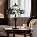 24.5" H Marlowe Multicolor Tiffany-Style Stained Glass Table Lamp sits on a round wooden side table in a living room setting next to a white coffee cup and a book, with a gray tufted sofa and curtains in the background.