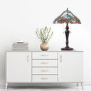24" H Highclere Victorian Multicolor Tiffany-Style Stained Glass Table Lamp on top of a white sideboard or credenza with five drawers and gold-tone handles, styled on a white floor against a white wall with a vase with greenery, and a stack of books.
